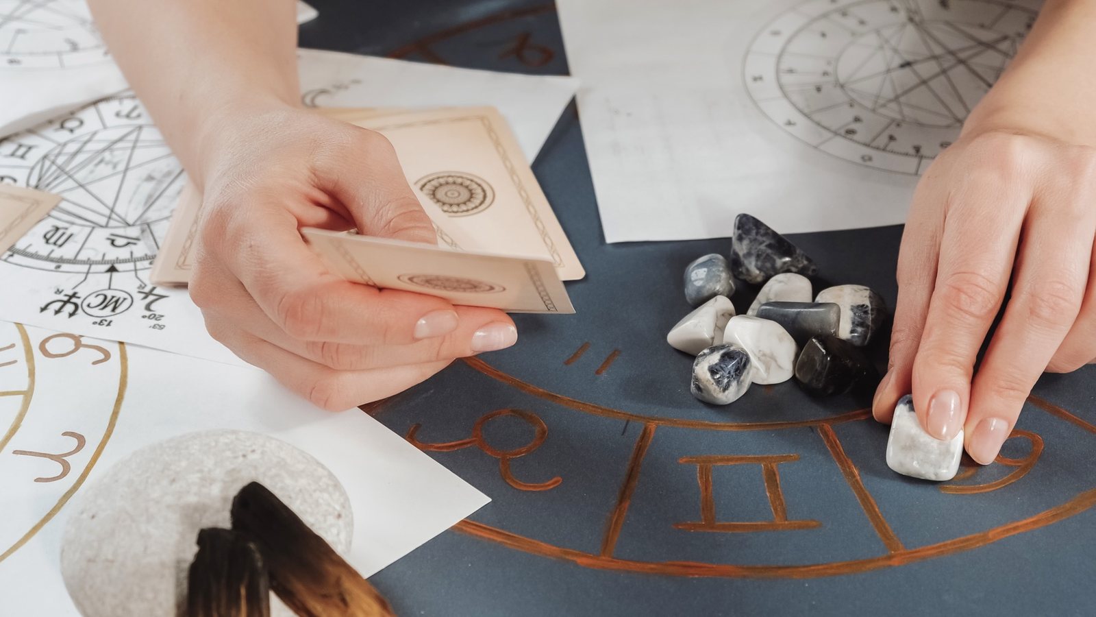 Mãos de uma astróloga sobre um mapa natal impresso, com pedras e cartas sobre a mesa