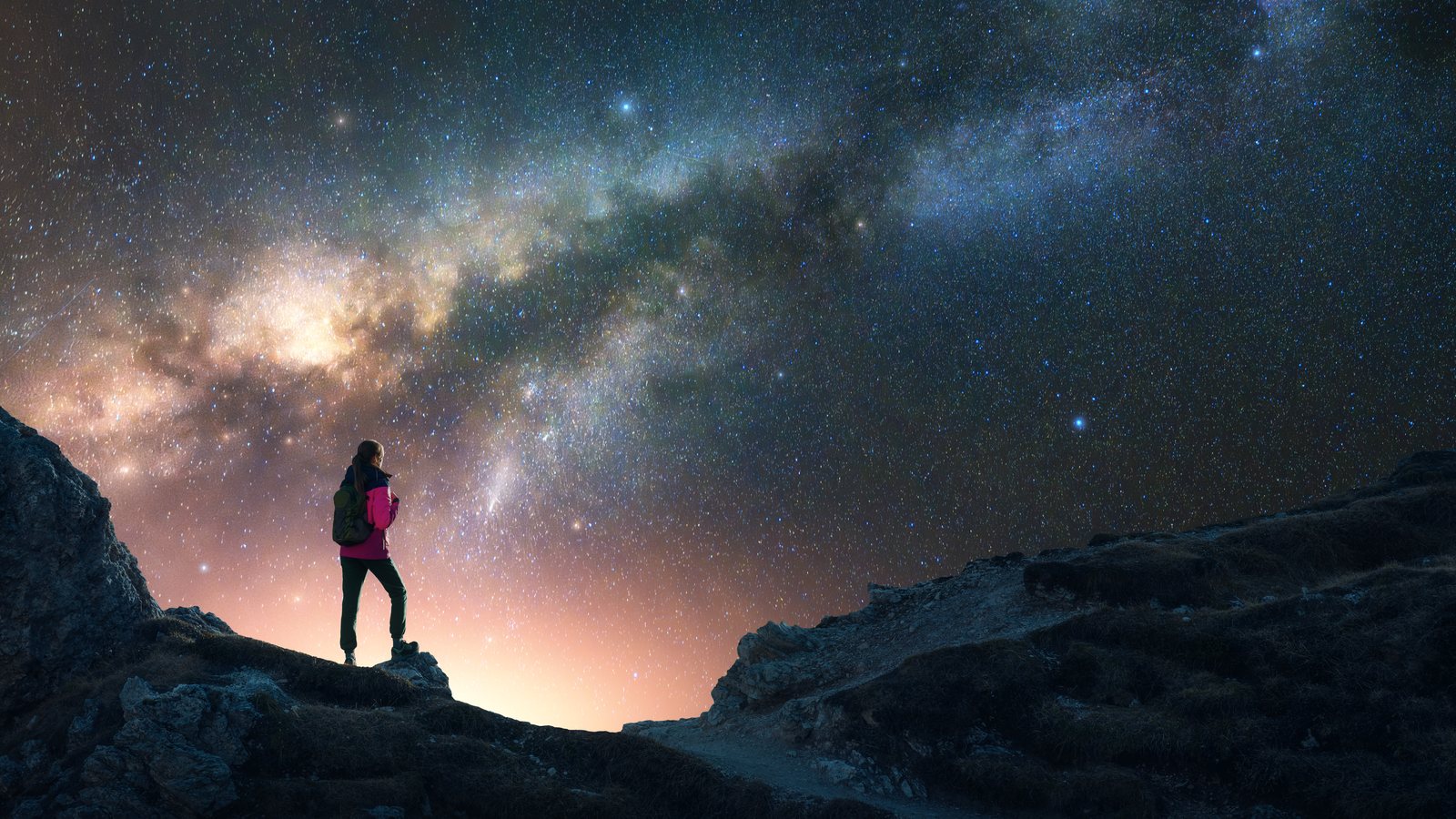 Mulher de costas no alto de uma montanha olhando a Via Láctea num céu noturno cheio de estrelas