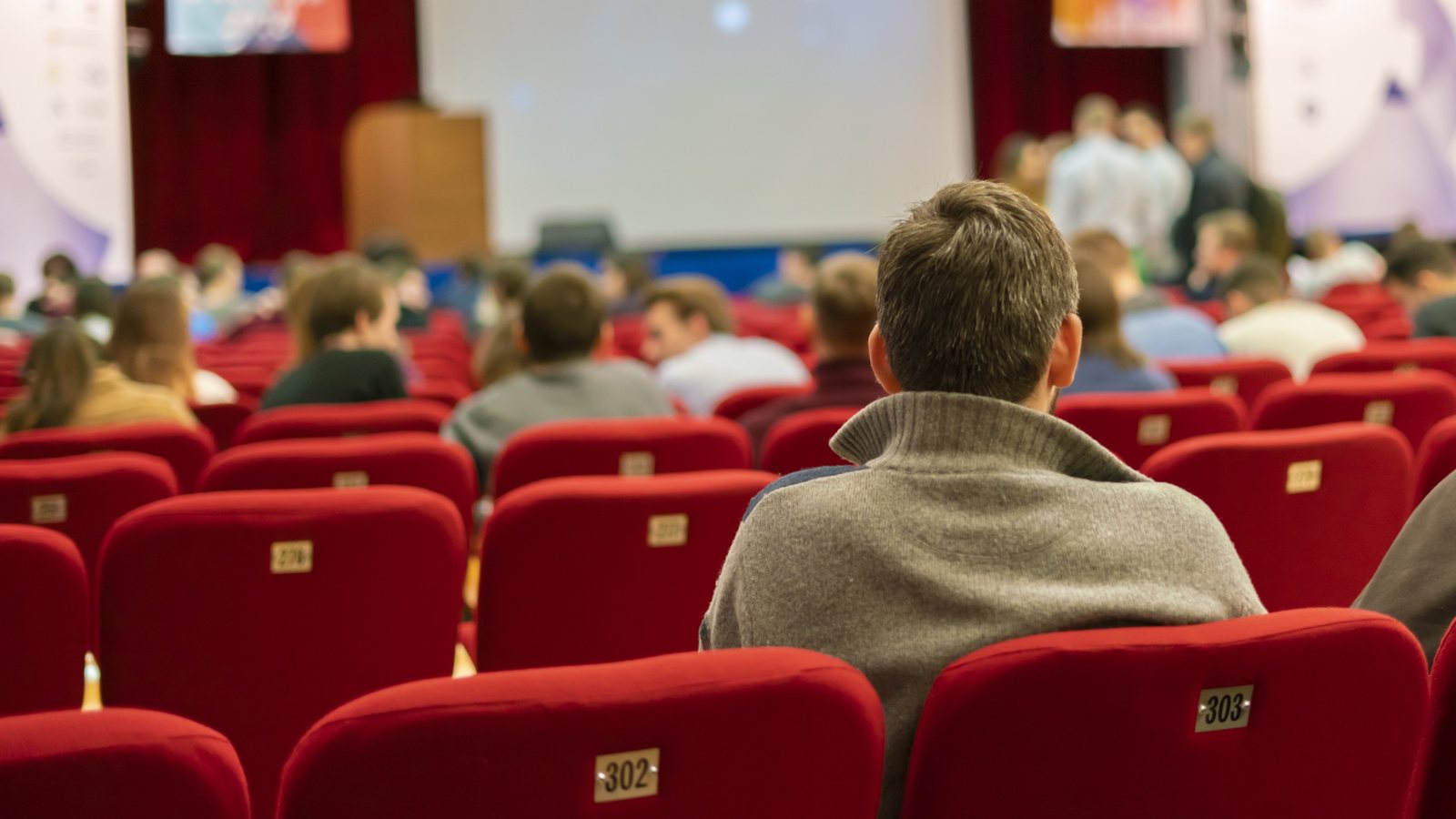 Plateia num auditório de poltronas vermelhas assistindo a uma palestra