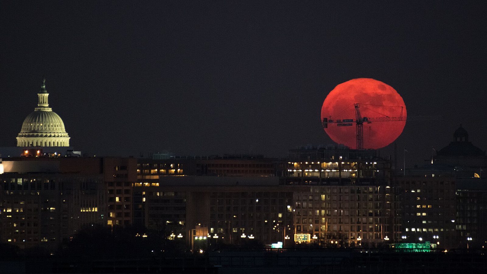 Lua cheia grande e alaranjada nascendo atrás de prédios iluminados de uma cidade à noite, como numa superlua