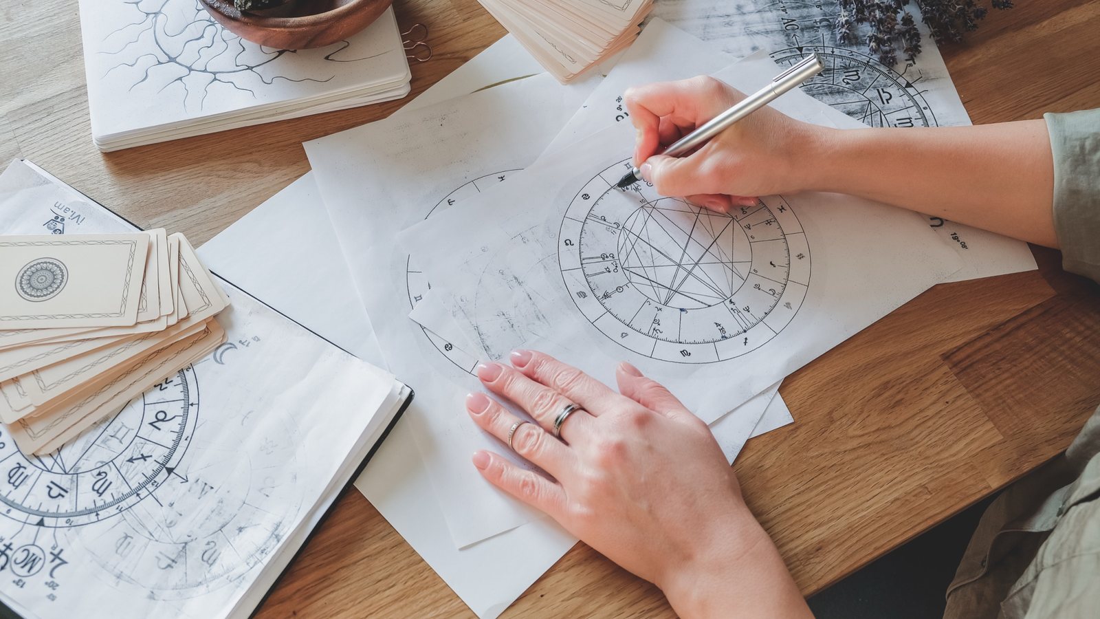 Mãos de uma astróloga desenham um mapa natal à mão sobre uma mesa de madeira, com cartas e anotações ao redor