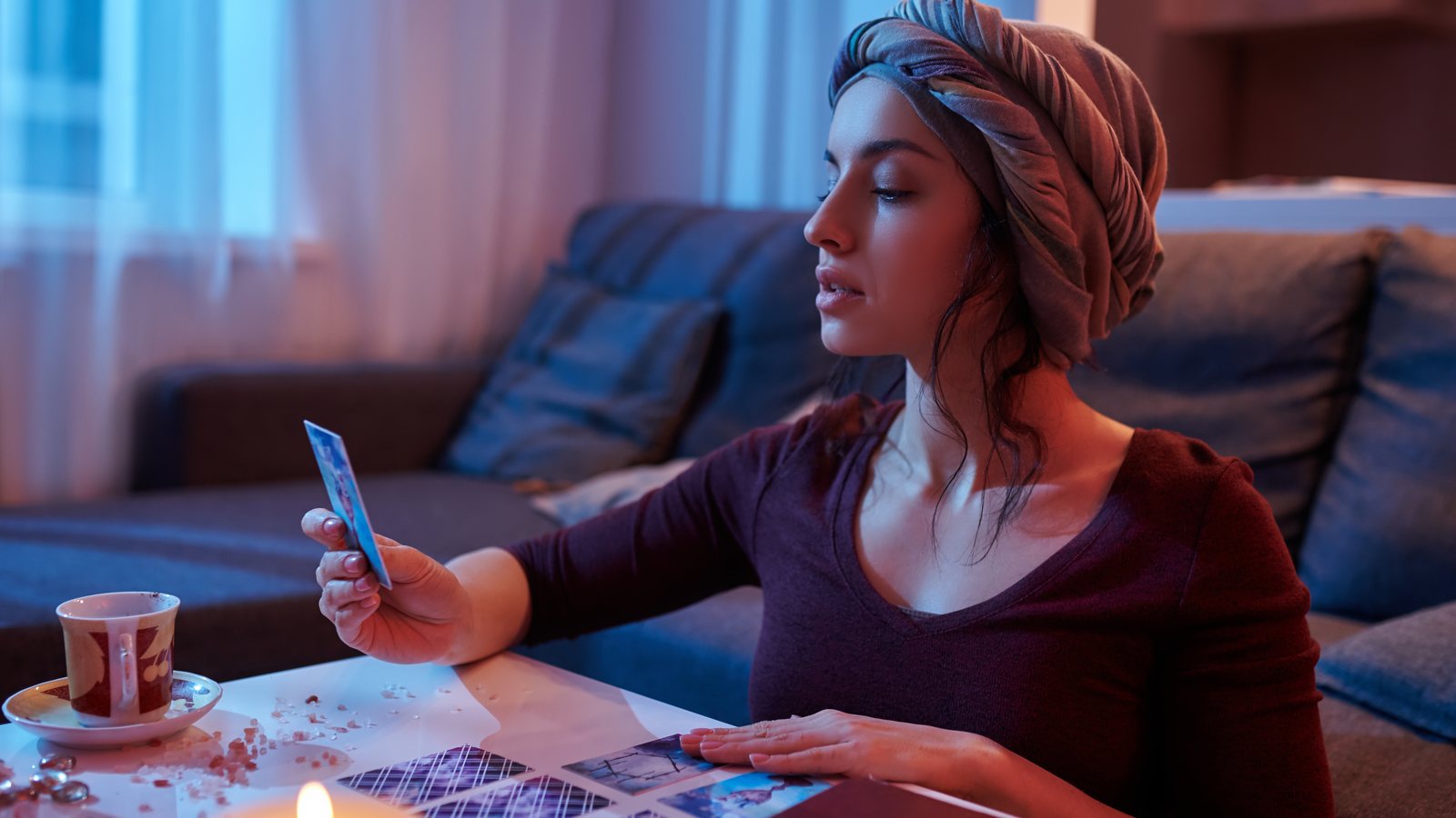 Jovem mulher sentada na sala olha uma carta de tarô, com outras cartas, uma xícara e uma vela sobre a mesa