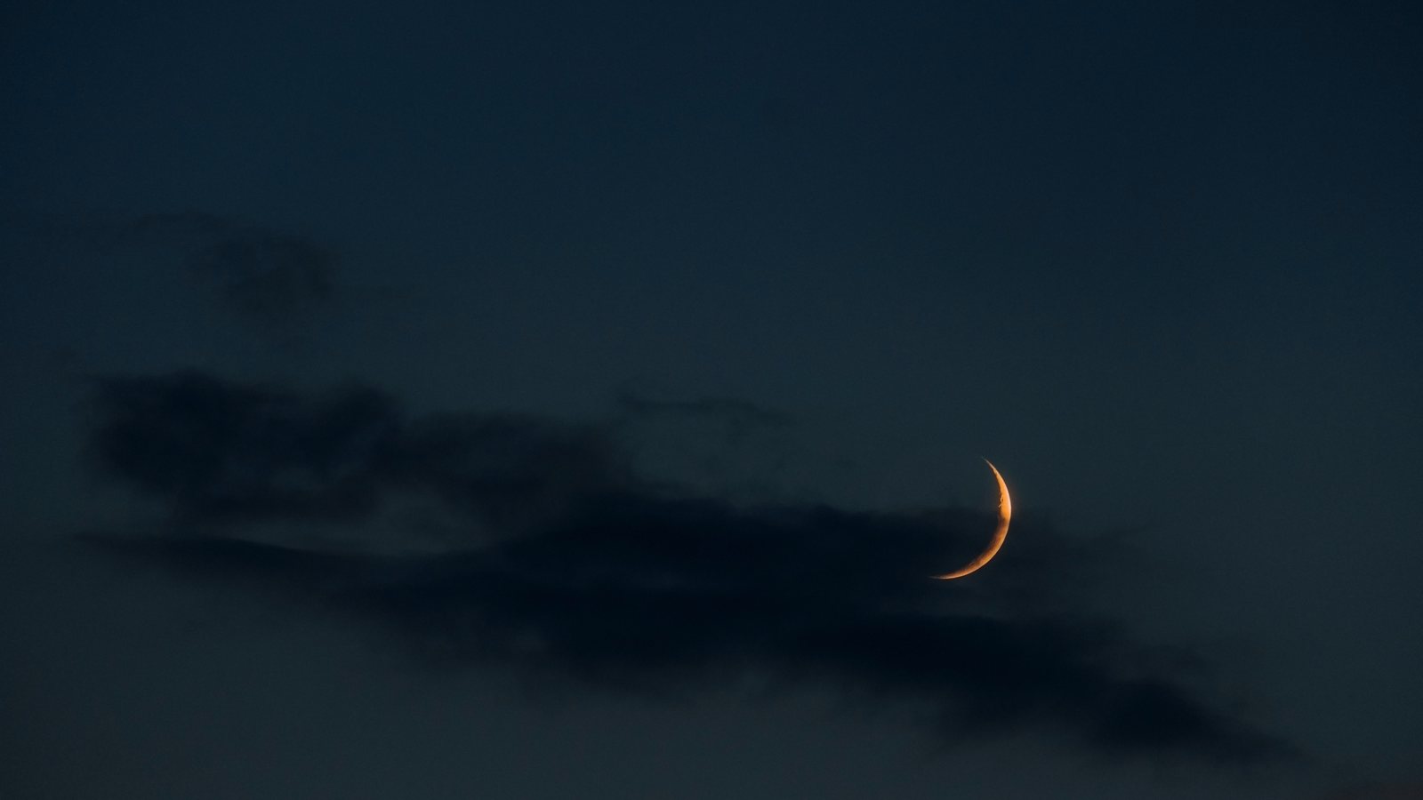 Lua crescente bem fina no céu azul escuro do entardecer, entre nuvens, como aparece um ou dois dias depois da Lua Nova