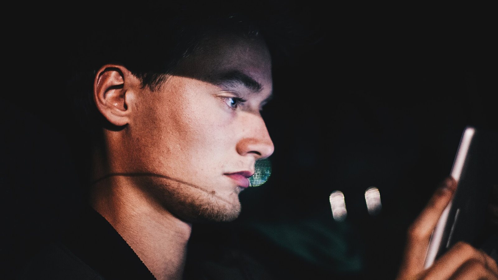 Jovem de perfil, no escuro, com o rosto iluminado pela tela do celular que segura na mão