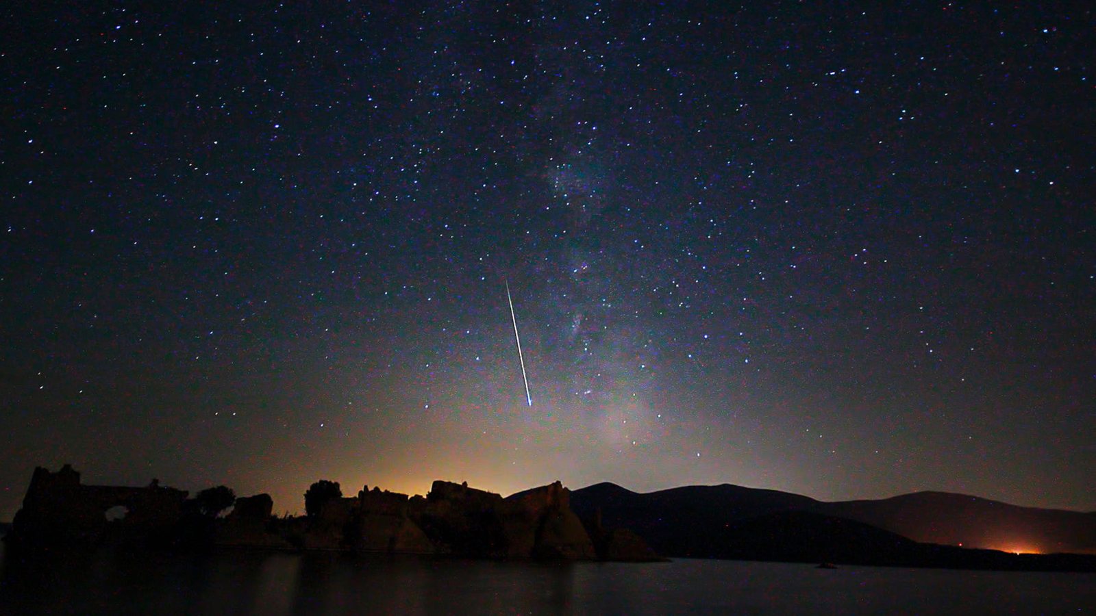 Meteoro risca o céu estrelado sobre um lago, com a Via Láctea ao fundo e o brilho de uma cidade no horizonte
