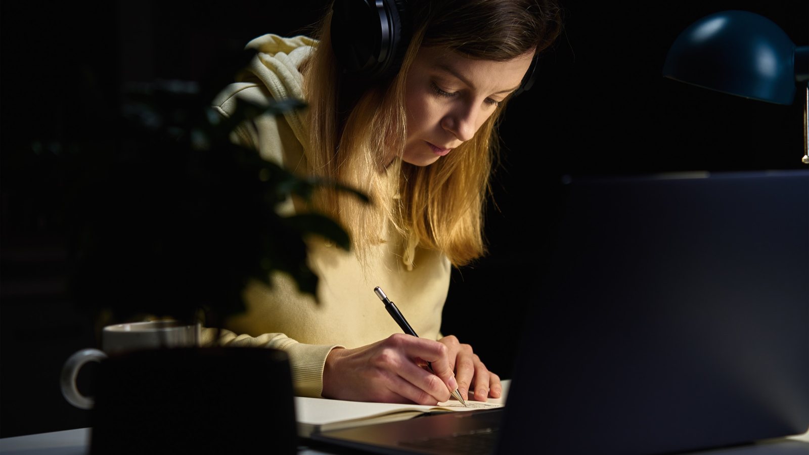 Mulher de fones de ouvido fazendo anotações à noite diante do notebook