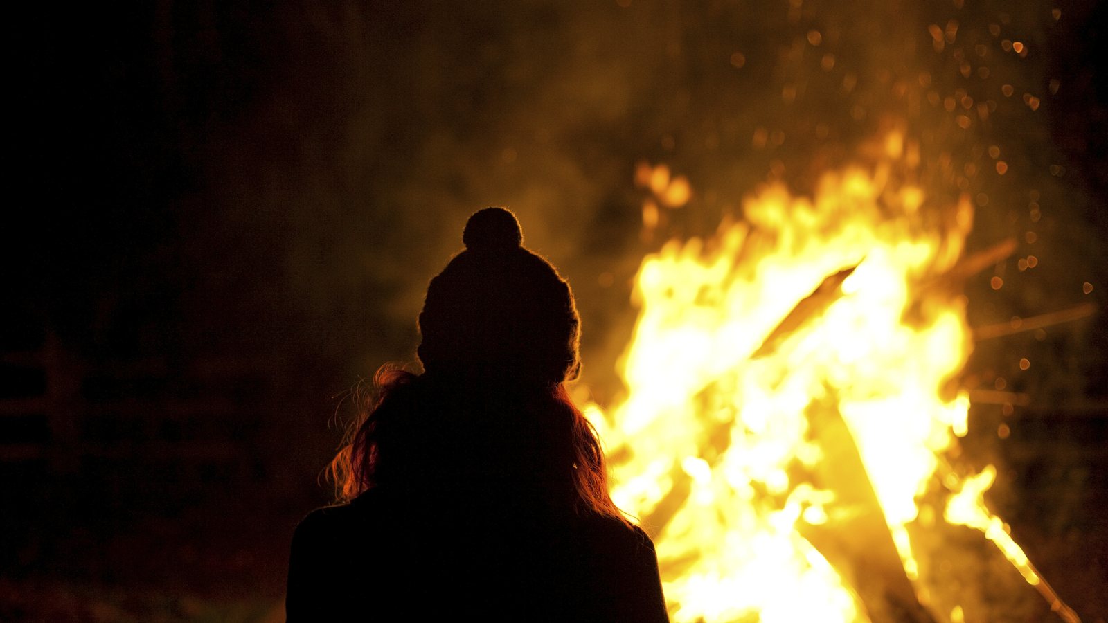 Silhueta de uma pessoa de gorro diante de uma grande fogueira acesa à noite