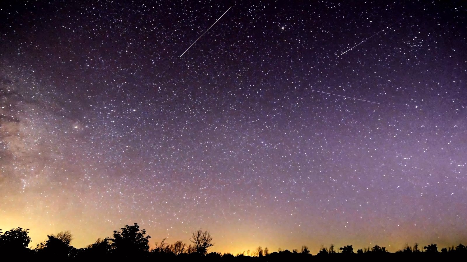 Céu estrelado de tom violeta sobre uma linha de árvores, com rastros de meteoros cortando a noite
